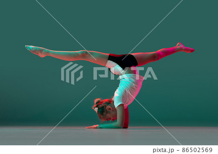 Handstand. Little flexible girl, rhythmic gymnastics artist training isolated on green studio background in neon pink light. Grace in motion, action. Doing exercises in flexibility. 86502569