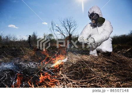 Unrecognizable person burning old dried grass in field. Focus on a fire. Ecologist holding matchbox and setting fire to dry grass under blue sky, wearing protective radiation suit and respirator. Unrecognizable person burning old dried grass in field. Focus on a fire. Ecologist holding matchbox and setting fire to dry grass under blue sky, wearing protective radiation suit and respirator. 86507148