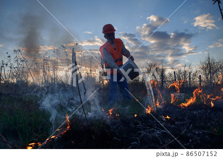 Fireman ecologist extinguishing fire in field with evening sky on background. Male environmentalist holding bucket and pouring water on burning dry grass. Natural disaster concept. Fireman ecologist extinguishing fire in field with evening sky on background. Male environmentalist holding bucket and pouring water on burning dry grass. Natural disaster concept. 86507152
