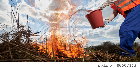 Fireman ecologist extinguishing fire in field with cloudy sky on background. Close up of man holding bucket and pouring water on burning dry grass. Natural disaster and ecology demage concept. Fireman ecologist extinguishing fire in field with cloudy sky on background. Close up of man holding bucket and pouring water on burning dry grass. Natural disaster and ecology demage concept. 86507153