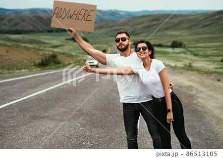 A couple in love hitchhiking. A young couple votes on the road. Travel on your own. Man and woman are hitchhiking. Couple Standing near Road. Man and woman catch a car on the road A couple in love hitchhiking. A young couple votes on the road. Travel on your own. Man and woman are hitchhiking. Couple Standing near Road. Man and woman catch a car on the road 86513105