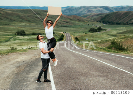 A young couple is voting on the road. A happy couple hitchhiking and voting with a sign on the road. Happy man and woman backpackers hikers travel together, vote on the road. Copy space A young couple is voting on the road. A happy couple hitchhiking and voting with a sign on the road. Happy man and woman backpackers hikers travel together, vote on the road. Copy space 86513109