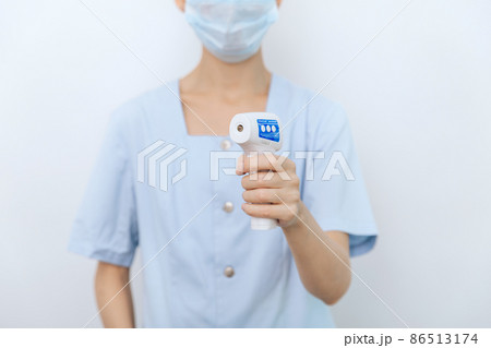 Temperature measurement gun in doctor hands. Close-up shot of doctor wearing protective surgical mask ready to use infrared isometric thermometer gun to check body temperature for virus symptoms. Temperature measurement gun in doctor hands. Close-up shot of doctor wearing protective surgical mask ready to use infrared isometric thermometer gun to check body temperature for virus symptoms. 86513174