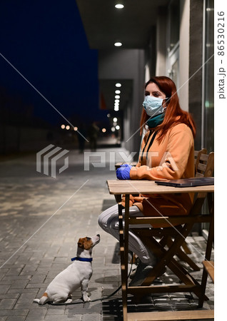 A woman in a medical mask and gloves walks late at night with her little dog. A girl sits on a chair in the street with a Jack Russell Terrier in quarantine. Coronavirus. 86530216