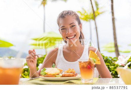 Happy woman eating lunch at outdoor restaurant terrace plant based meat burger with pineapple. Hawaii japanese tropical food. Asian girl at hotel cafe table during summer travel vacation Happy woman eating lunch at outdoor restaurant terrace plant based meat burger with pineapple. Hawaii japanese tropical food. Asian girl at hotel cafe table during summer travel vacation 86537791