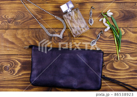 Women accessories on wooden background. Clutch bag, bottle of perfume, necklace, earrings and snowdrops on wood table. Beauty and fashion composition. Top view, flat lay 86538923