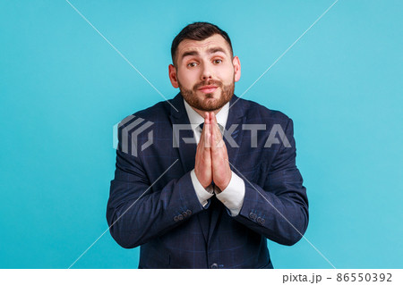 Please, I'm begging. Unhappy bearded man in official style suit keeping hands in prayer looking with imploring expression, sincere asking permission. Indoor studio shot isolated on blue background. Please, I'm begging. Unhappy bearded man in official style suit keeping hands in prayer looking with imploring expression, sincere asking permission. Indoor studio shot isolated on blue background. 86550392