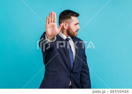Stop. keep your distance. Serious bearded man wearing official style suit standing with stop hand and looking away, showing ban, block sign with hand. Indoor studio shot isolated on blue background. 86550488