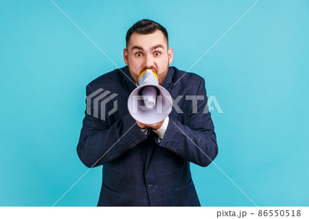 Portrait of angry nervous bearded man wearing elegant suit loudly screaming at megaphone, making announce, protesting, wants to be heard. Indoor studio shot isolated on blue background. Portrait of angry nervous bearded man wearing elegant suit loudly screaming at megaphone, making announce, protesting, wants to be heard. Indoor studio shot isolated on blue background. 86550518