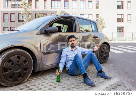 Young adult upset driver man wearing jeans and blue shirt sitting near crash car, collision accident in city road, dont drive drunk, damaged car door with dents and scratches. Outdoor shot. Young adult upset driver man wearing jeans and blue shirt sitting near crash car, collision accident in city road, dont drive drunk, damaged car door with dents and scratches. Outdoor shot. 86550757