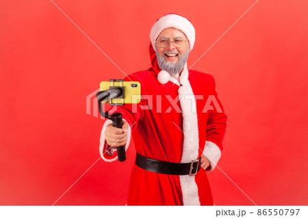 Attractive elderly man with gray beard wearing santa claus costume holding steadicam, broadcasting livestream, blogger talking with followers. Indoor studio shot isolated on red background. Attractive elderly man with gray beard wearing santa claus costume holding steadicam, broadcasting livestream, blogger talking with followers. Indoor studio shot isolated on red background. 86550797