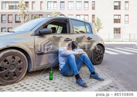 Man wearing jeans and blue shirt sitting near crashed car with bottle of alcohol beverage and sleeping, damaged automobile drunk driving, violation led to road accident. Outdoor shot. 86550806