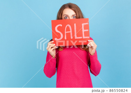 Portrait of curious woman wearing pink pullover, peeping from red card with sale inscription, mega shopping, black friday discounts. Indoor studio shot isolated on blue background. 86550824