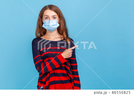 Portrait of serious woman wearing striped casual style sweater and surgical mask pointing finger side for empty wall for advertisement. Indoor studio shot isolated on blue background. 86550825