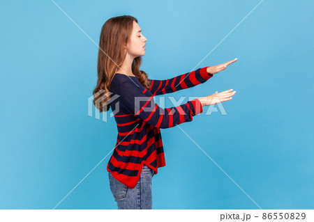 Side view portrait of blind woman in striped casual style sweater, walking alone with closed eyes in darkness, feeling disoriented confused, lost road. Indoor studio shot isolated on blue background. 86550829
