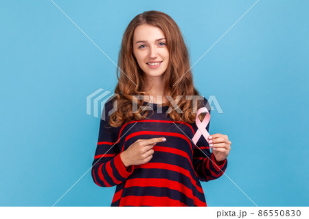 Woman wearing striped casual style sweater, points at pink ribbon, symbol of breast cancer awareness, female healthcare, medical insurance. Indoor studio shot isolated on blue background. 86550830