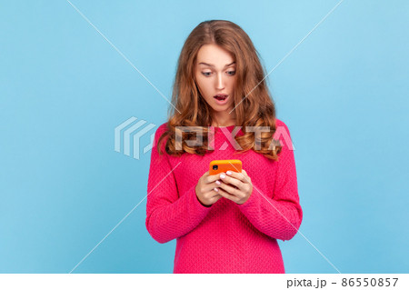 Portrait of amazed woman wearing pink pullover, looking at cellphone with surprised expression, reading shocking news using mobile phone. Indoor studio shot isolated on blue background. Portrait of amazed woman wearing pink pullover, looking at cellphone with surprised expression, reading shocking news using mobile phone. Indoor studio shot isolated on blue background. 86550857