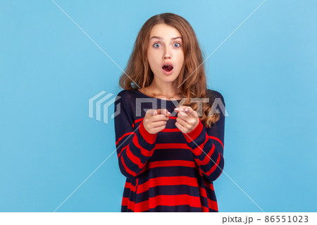 Astonished woman with wavy hair wearing striped casual style sweater pointing to camera with surprised shocked expression, making choice. Indoor studio shot isolated on blue background. 86551023