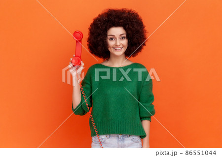 Portrait of smiling woman with curly hair wearing green casual style sweater standing holding red retro land line phone, waiting conversation. Indoor studio shot isolated on orange background. Portrait of smiling woman with curly hair wearing green casual style sweater standing holding red retro land line phone, waiting conversation. Indoor studio shot isolated on orange background. 86551044