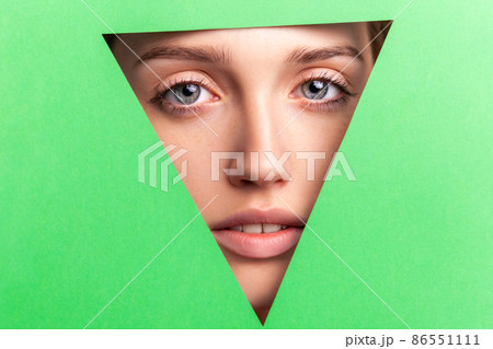 Closeup portrait of well-groomed woman with natural makeup, looking at camera through triangular hole of green paper, beauty and health and skin care concept. Indoor studio shot. 86551111