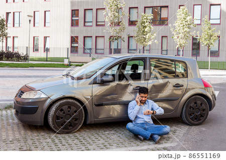 Young adult bearded man wearing jeans and shirt sitting on ground, looking at wristwatch and talking phone, waiting insurance inspector, broken car with dents and scratches. Outdoor shot. 86551169