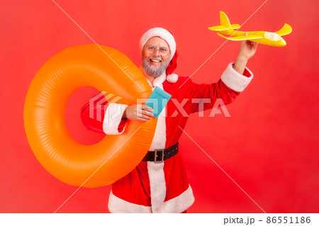 Happy elderly man with gray beard wearing santa claus costume holding orange rubber ring, paper plane and passport, traveling during winter holidays. Indoor studio shot isolated on red background. Happy elderly man with gray beard wearing santa claus costume holding orange rubber ring, paper plane and passport, traveling during winter holidays. Indoor studio shot isolated on red background. 86551186