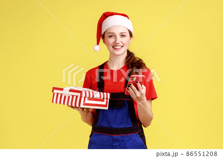 Good looking delivery woman holding wrapped present box and using phone, communicate with client, wearing blue overalls and santa claus hat. Indoor studio shot isolated on yellow background. 86551208