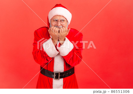 Portrait of elderly man with gray beard wearing santa claus costume biting nails, being nervous terrified, feeling frightened, anxiety disorder. Indoor studio shot isolated on red background. 86551306