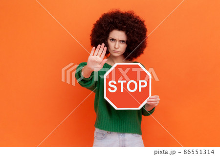 Serious woman with curly hair wearing green casual style sweater showing stop gesture with hand holding red traffic sign of prohibition, blocking. Indoor studio shot isolated on orange background. 86551314