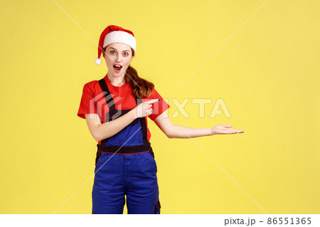 Female worker presenting copy space on palm, points finger aside, advertisement of service industry, wearing blue overalls and santa claus hat. Indoor studio shot isolated on yellow background. 86551365