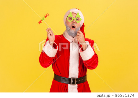 Excited elderly man with gray beard wearing santa claus costume holding paper cards on sticks, celebrating christmas and congratulating with new year. Indoor studio shot isolated on yellow background. 86551374