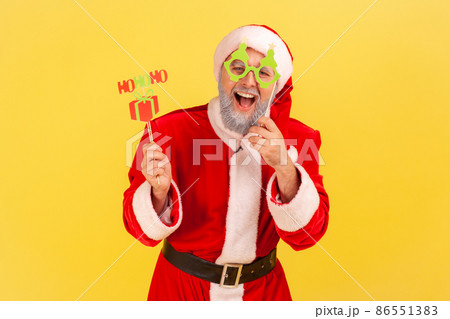 Excited elderly man with gray beard wearing santa claus costume holding congratulations cards on sticks, new year and christmas celebration. Indoor studio shot isolated on yellow background. Excited elderly man with gray beard wearing santa claus costume holding congratulations cards on sticks, new year and christmas celebration. Indoor studio shot isolated on yellow background. 86551383