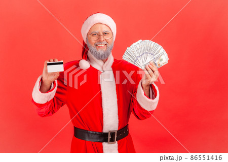 Satisfied elderly man with gray beard wearing santa claus costume standing holding fan of dollars and showing credit card, cashback. Indoor studio shot isolated on red background. 86551416