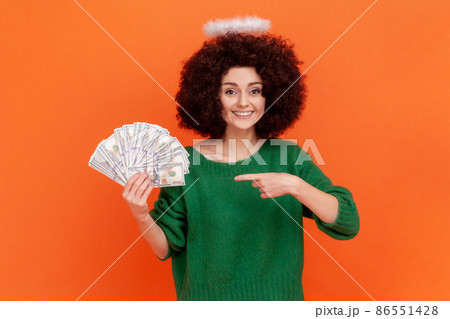 Young adult angelic woman with curly hair wearing green casual style sweater with nimb over head holding and pointing at fan of dollars banknotes. Indoor studio shot isolated on orange background. 86551428