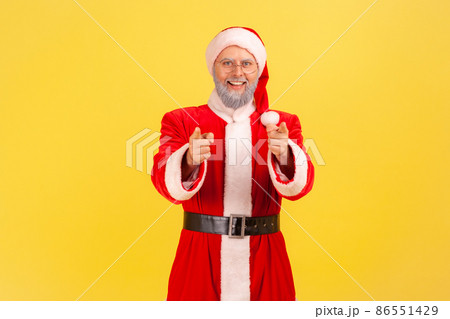 Pleasant looking positive elderly man with gray beard wearing santa claus costume standing pointing to camera with smile, choosing you. Indoor studio shot isolated on yellow background. Pleasant looking positive elderly man with gray beard wearing santa claus costume standing pointing to camera with smile, choosing you. Indoor studio shot isolated on yellow background. 86551429