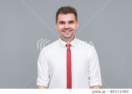 Portrait of young handsome happy successful man in white shirt and tie standing and looking at camera with toothy smile. indoor isolated on gray background. 86551460