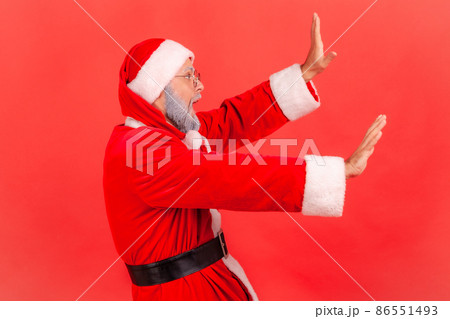 Profile portrait of elderly man with gray beard wearing santa claus costume raising hands from sudden fear, looking terrified, scared to death. Indoor studio shot isolated on red background. 86551493
