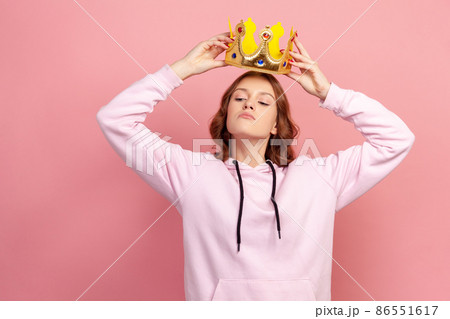 Portrait of selfish curly haired teenage girl in hoodie holding gold crown over head, looking with arrogance, privileged status. Indoor studio shot isolated on pink background 86551617