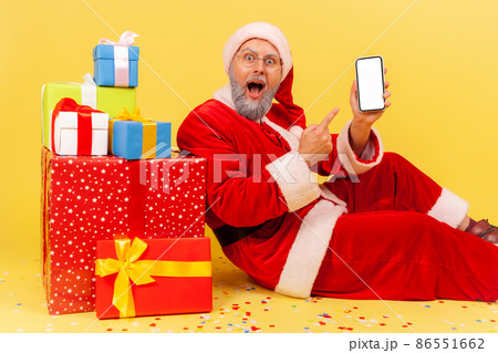 Astonished elderly man with gray beard wearing santa claus costume sitting pointing white blank screen of smart phone, surrounded with Christmas gifts. Indoor studio shot isolated on yellow background 86551662