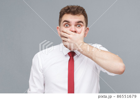 Portrait of young shocked man in white shirt and tie standing closing his mouth with hand and looking at camera with shocked face and big eyes. indoor isolated on gray background. Portrait of young shocked man in white shirt and tie standing closing his mouth with hand and looking at camera with shocked face and big eyes. indoor isolated on gray background. 86551679