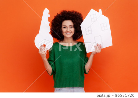 Good looking smiling woman with Afro hairstyle wearing green casual style sweater holding big paper key and house, expressing positive emotions. Indoor studio shot isolated on orange background. 86551720