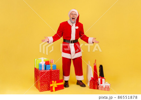 Full length portrait of elderly man with gray beard in santa claus costume standing with excited expression and raised arms near Christmas gifts. Indoor studio shot isolated on yellow background. Full length portrait of elderly man with gray beard in santa claus costume standing with excited expression and raised arms near Christmas gifts. Indoor studio shot isolated on yellow background. 86551878