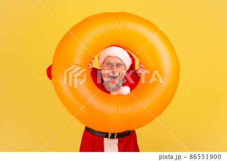 Portrait of elderly man with gray beard wearing santa claus costume has amazed shocked expression, celebrating winter holidays in warm resort. Indoor studio shot isolated on yellow background. 86551900
