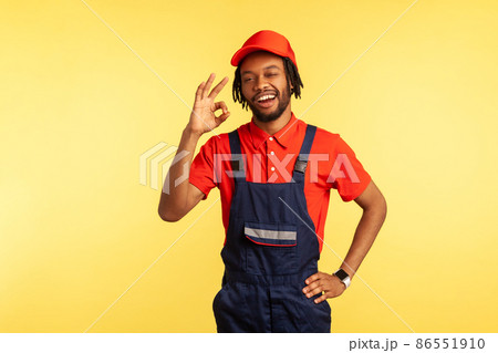 Portrait of sappy handyman with beard wearing overalls showing ok sign, being satisfied of done work in time, recommend service industry. Indoor studio shot isolated on yellow background. 86551910