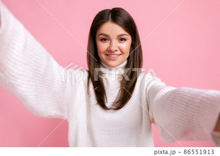Portrait of smiling happy brunette female taking selfie, point of view photo, expression optimism, wearing white casual style sweater. Indoor studio shot isolated on pink background. 86551913