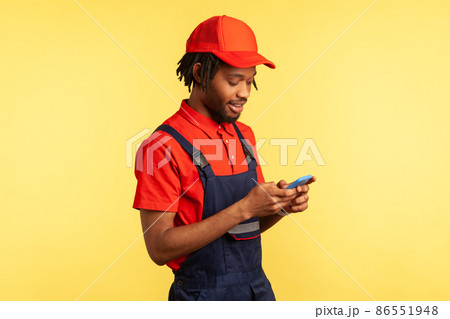 Side view portrait of positive deliveryman wearing uniform, using smartphone for taking new order for delivering, delivery service. Indoor studio shot isolated on yellow background. 86551948