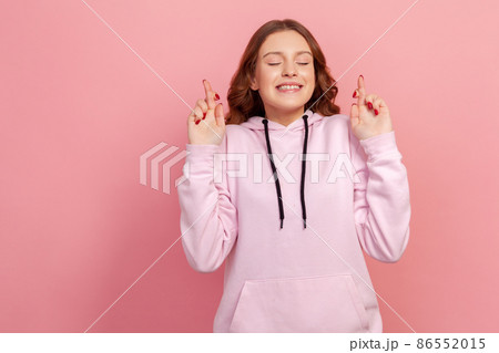 Portrait of hopeful joyous young woman with curly hair in hoodie raising fingers crossed while making wish, confident to win. Indoor studio shot isolated on pink background 86552015
