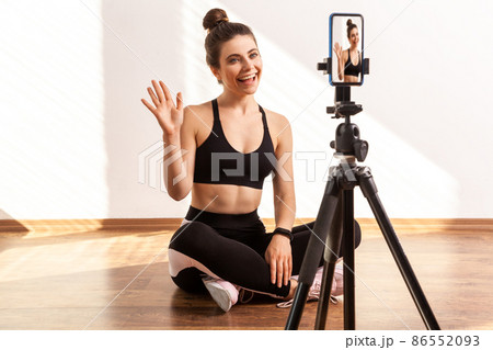 Trainer saying hello or bye, recording online tutorial of fitness exercises, using hone on tripod, wearing black sports top and tights. Full length studio shot illuminated by sunlight from window. Trainer saying hello or bye, recording online tutorial of fitness exercises, using hone on tripod, wearing black sports top and tights. Full length studio shot illuminated by sunlight from window. 86552093