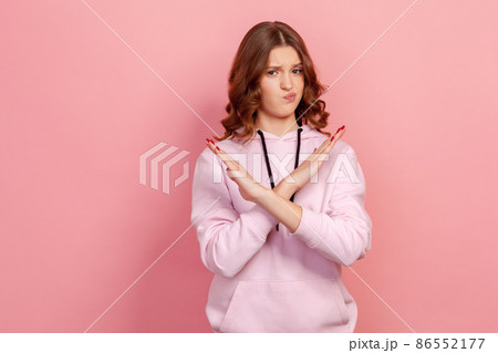 Portrait of serious teen girl with brunette hair in hoodie making x sign with crossed hands, gesturing stop, warning to ban, body language. Indoor studio shot isolated on pink background 86552177