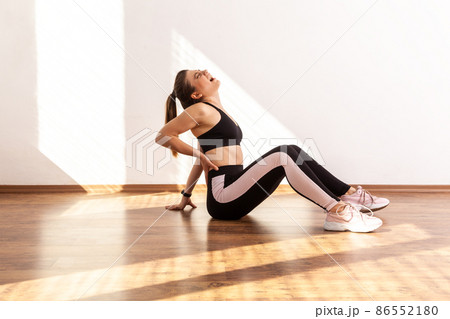 Side view of slim girl sitting touching sore spine after workout, has trauma, feeling discomfort, wearing black sports top and tights. Full length studio shot illuminated by sunlight from window. 86552180